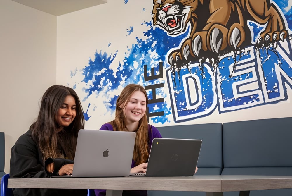 Zwei Schülerinnen der Covenant Christian School sitzen an einem Tisch und arbeiten an Laptops vor einem Tiger-Wandbild.
