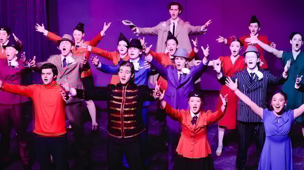 Schüler der Covenant Day School führen eine lebhafte Bühnenaufführung in bunten Kostümen mit erhobenen Armen auf.