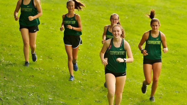 Eine Gruppe junger Athletinnen der Coventry High School läuft auf einem grasbewachsenen Feld bei Sonnenschein.