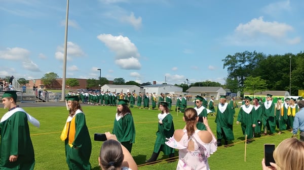 Absolventinnen und Absolventen der Coventry High School in grünen und gelben Talaren stehen auf einer Wiese vor einem Backsteingebäude.