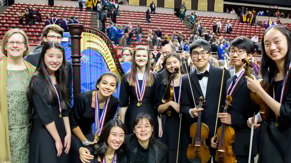 Eine Gruppe junger Musikerinnen und Musiker posiert auf der Bühne des Cranbrook Education Campus vor roten Sitzen und einer großen Harfe.