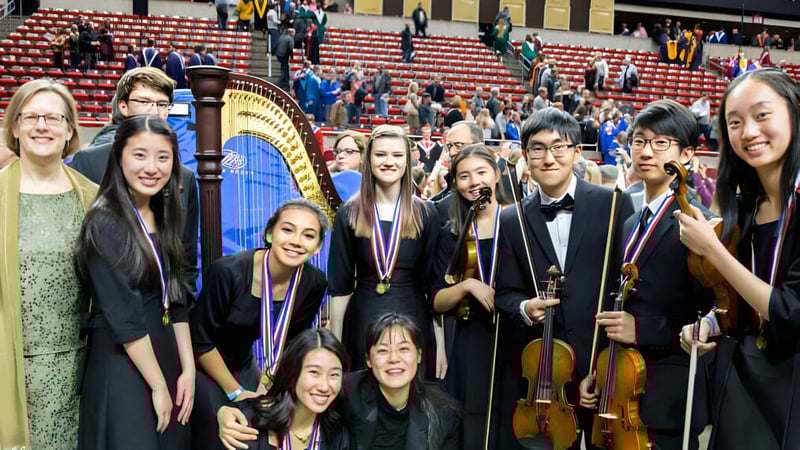 Eine Gruppe junger Musikerinnen und Musiker posiert auf der Bühne des Cranbrook Education Campus vor roten Sitzen und einer großen Harfe.