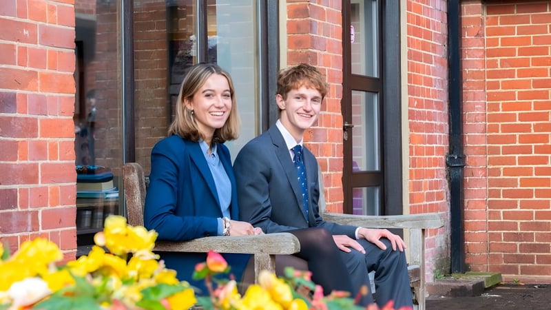 Zwei Schülerinnen und Schüler der Cranleigh School sitzen zusammen auf einer Bank vor einem Backsteingebäude mit gelben Blumen.
