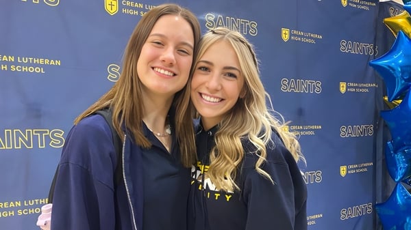 Zwei lächelnde Schülerinnen mit langen blonden Haaren stehen vor einem Hintergrund mit dem Logo der Crean Lutheran High School.