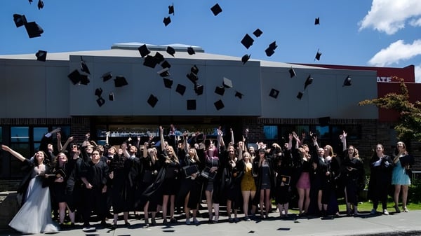 Eine große Gruppe von Graduierenden der Crescent Collegiate wirft ihre Abschlusskappen vor einem Gebäude in die Luft.
