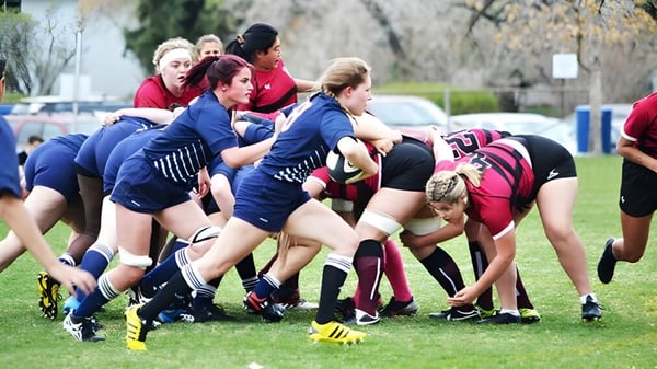 Schülerinnen der Crescent Heights High School befinden sich bei einem Rugby-Scrum auf einem Rasenfeld mit Bäumen und Gebäuden im Hintergrund.