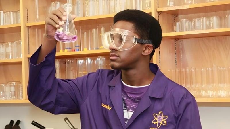 Ein Schüler der Crescent Heights High School hält in einem Labor einen Becher mit pinker Flüssigkeit vor einem Bücherregal hoch.