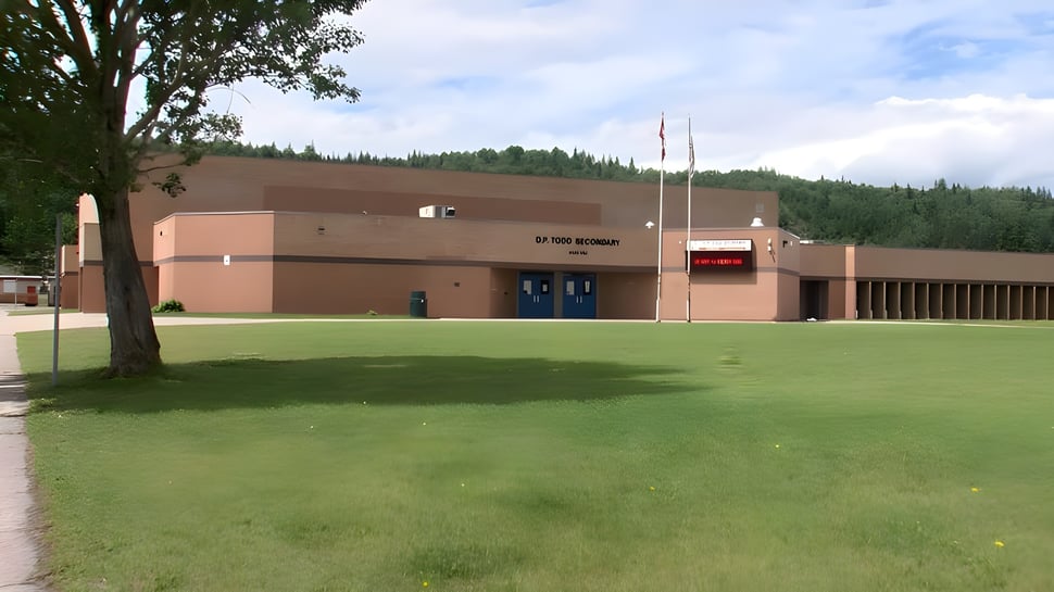 Das große braune Schulgebäude der D.P. Todd Secondary School steht auf einer Wiese umgeben von Bäumen unter bewölktem Himmel.