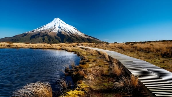 Ein Holzsteg führt durch eine Wiesenlandschaft zu einem schneebedeckten Berg, der sich im See nahe der Dannevirke High School spiegelt.