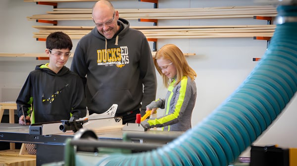 Schüler der École Dansereau Meadows School arbeiten gemeinsam mit einem Erwachsenen in einer Werkstatt mit Werkzeugen im Hintergrund.