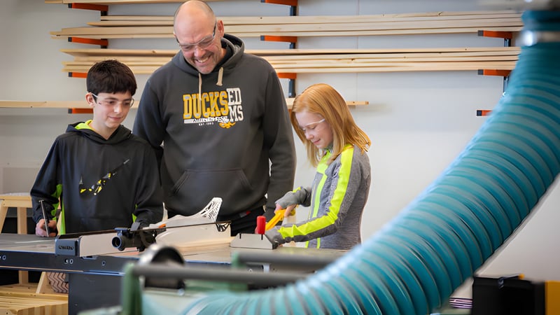 Schüler der École Dansereau Meadows School arbeiten gemeinsam mit einem Erwachsenen in einer Werkstatt mit Werkzeugen im Hintergrund.