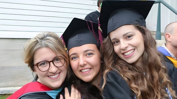 Drei junge Frauen in Abschlusskleidung der Darrow School stehen vor einem Holzgebäude.