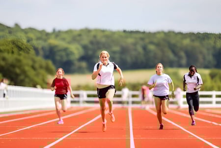 Eine Gruppe Läuferinnen sprintet auf der roten Laufbahn auf dem Campus der Dauntsey’s School vor bewaldeter Landschaft.