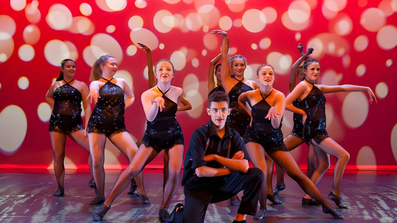 Eine Gruppe von Tänzerinnen und Tänzern der Davidson High School führt auf einer Bühne vor rotem Hintergrund mit weißen Punkten auf.