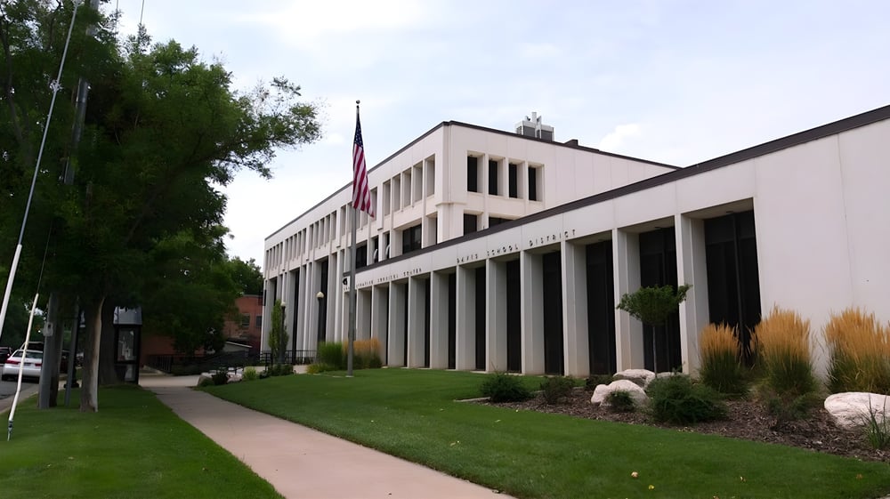 Das moderne mehrstöckige Gebäude des Davis School District mit Fahnenmast und gepflegten Grünflächen umgeben von Bäumen.