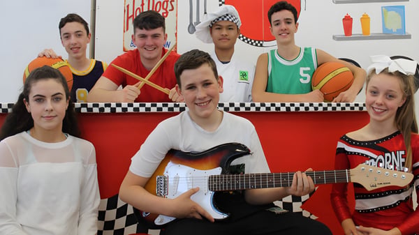 Eine Gruppe von Schülerinnen und Schülern mit Musikinstrumenten und Sportgeräten vor einem bunten karierungsmuster auf dem Campus des Davitt College.