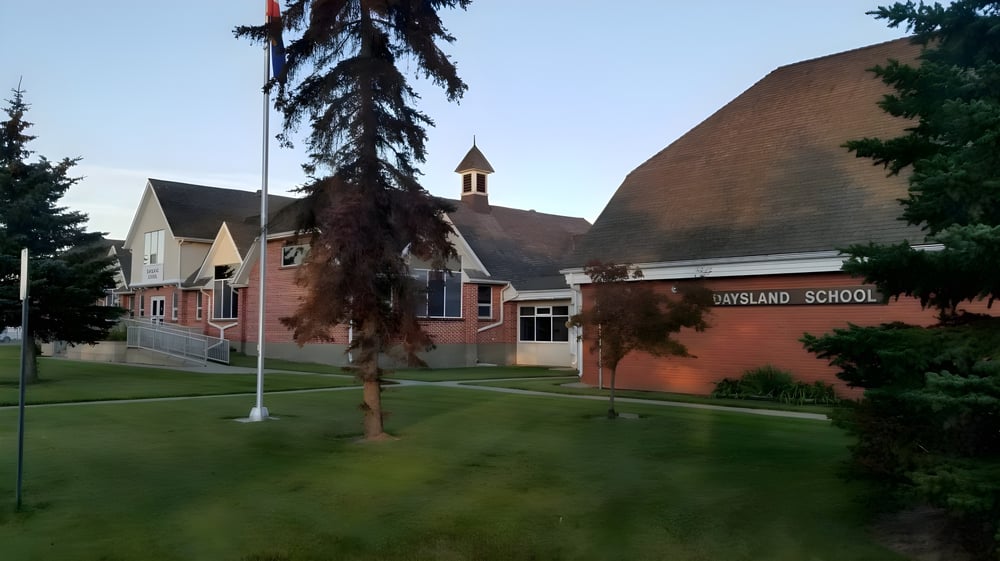 Das Backsteingebäude der Daysland School mit Glockenturm steht auf einer grasbewachsenen Fläche umgeben von Bäumen unter blauem Himmel.