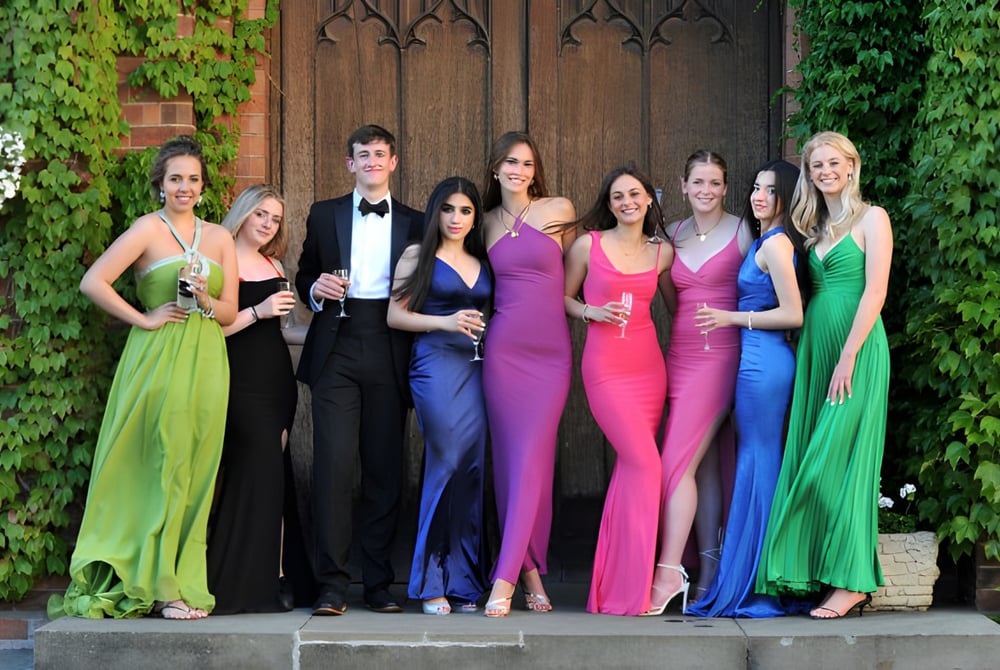 Eine Gruppe elegant gekleideter Personen posiert gemeinsam vor einer Holztür umgeben von grünen Pflanzen auf dem Gelände der Dean Close School.