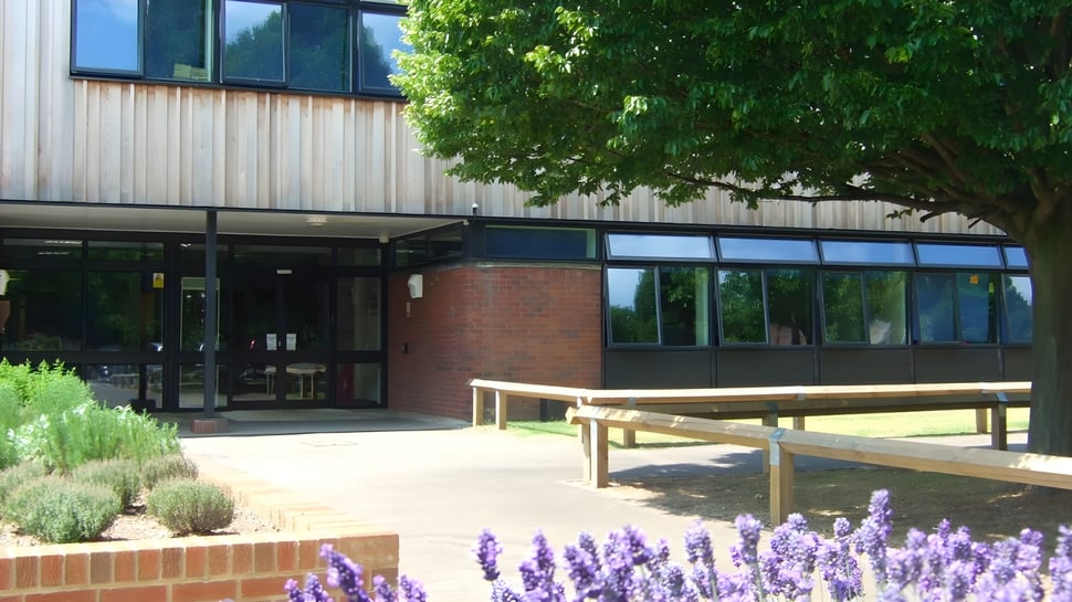 Das moderne Schulgebäude der Denefield School ist von grüner Bepflanzung mit lila Blumen und einer Holzbank umgeben.