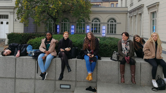Eine Gruppe von Schülerinnen der Denefield School sitzt auf einer Steinmauer vor einem historischen Gebäude mit Bögen.