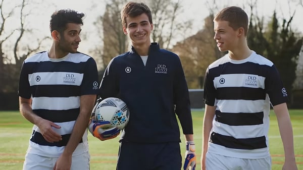 Drei Schüler des DLD College London stehen gemeinsam auf einem Rasenfeld mit Bäumen und bewölktem Himmel im Hintergrund.