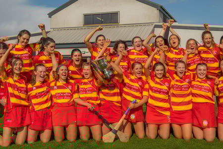 Eine Gruppe junger Sportlerinnen des Dominican College Galway feiert gemeinsam auf einem Sportfeld vor einem Gebäude.