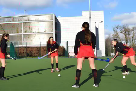 Schülerinnen des Dominican College Sion Hill spielen Feldhockey auf dem Kunstrasenplatz mit einem Schulgebäude im Hintergrund.