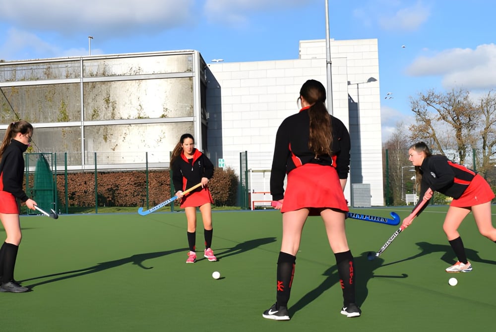 Schülerinnen des Dominican College Sion Hill spielen Feldhockey auf dem Kunstrasenplatz mit einem Schulgebäude im Hintergrund.