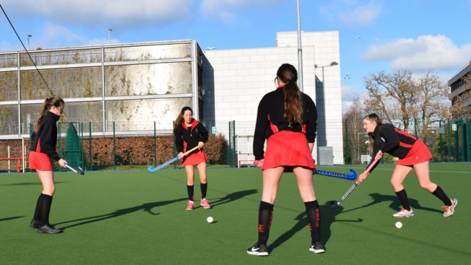 Schülerinnen des Dominican College Sion Hill spielen Feldhockey auf einem Kunstrasenfeld mit Gebäude im Hintergrund.