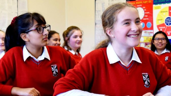 Schüler des Dominican College Sion Hill stehen in roten Schuluniformen in einem Klassenraum mit Lernpostern im Hintergrund.