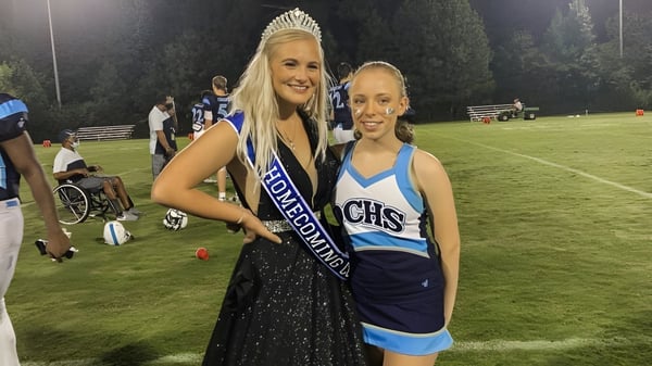 Zwei Schülerinnen der Dominion Christian School stehen bei Nacht auf dem Sportfeld, eine als Homecoming Queen mit Krone und Schärpe, die andere im Cheerleader-Uniform.