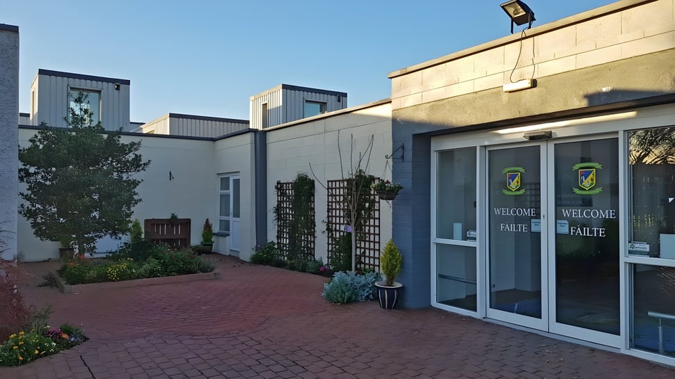 Der gepflasterte Eingangsbereich führt zu dem modernen Gebäude der Donahies Community School mit Glasfront und Pflanzen.