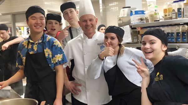 Schüler der Dover Bay Secondary School versammeln sich in der Schulküche mit einem Koch in weißer Uniform.