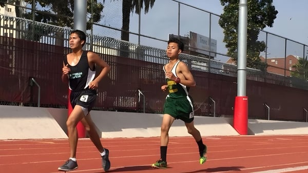 Zwei Athleten der Downtown Magnet High School laufen auf der roten Sportbahn mit Zaun und Gebäuden im Hintergrund.