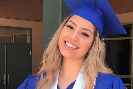 Eine junge Frau mit blauer Abschlussmütze und -robe steht lächelnd vor einer Glastür auf dem Campus der Dr. Charles Best Secondary School.