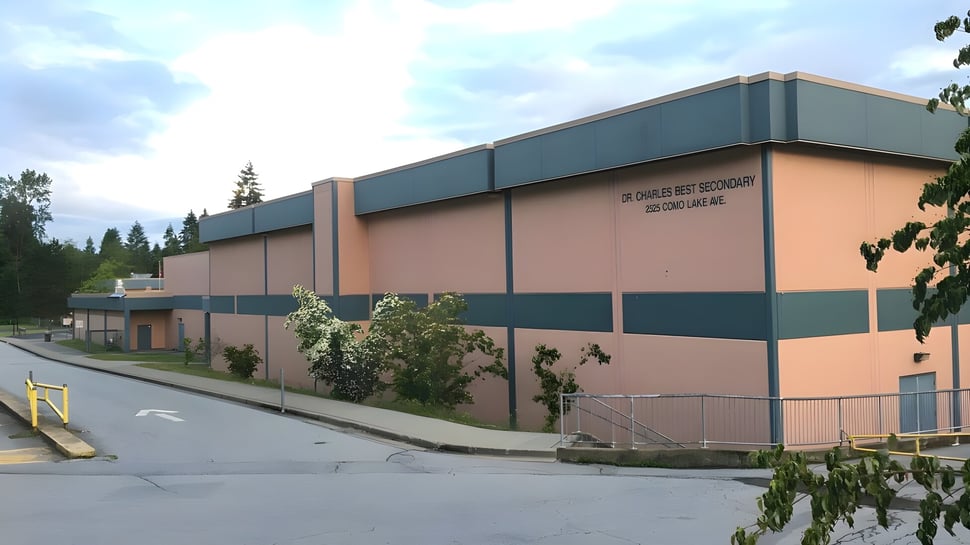 Das mehrstöckige Gebäude der Dr. Charles Best Secondary School mit rosa Fassade und grünen Akzenten steht von Bäumen umgeben.
