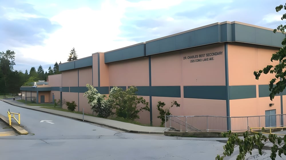 Das mehrstöckige Gebäude der Dr. Charles Best Secondary School mit rosa Fassade und grünen Akzenten steht von Bäumen umgeben.