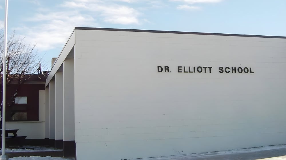 Das moderne Schulgebäude der Dr. Elliott School mit deutlich sichtbarem Schriftzug unter bewölktem Himmel.