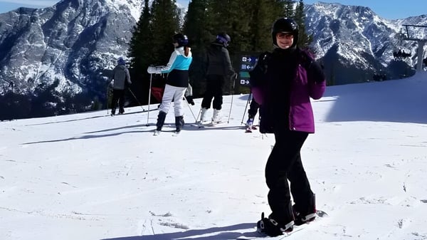 Eine Schülerin der Dr. Elliott School trägt Ski-Ausrüstung auf einer verschneiten Piste mit Bergen und Kiefern im Hintergrund.