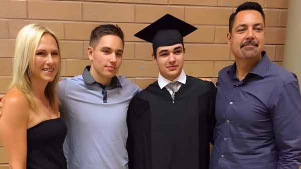 Eine Gruppe von vier Personen, darunter ein Schüler in einer Abschlussrobe, steht vor einer Backsteinwand auf dem Gelände der Dr. Frank J. Hayden Secondary School.