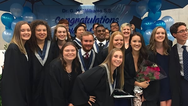 Schüler der Dr. G.W. Williams Secondary School versammeln sich in Abschlussgewändern vor einem Glückwunschbanner mit blauem Regenschirm darüber.
