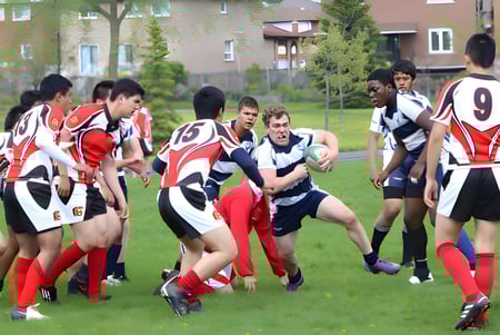 Schüler des Dr. Norman Bethune Collegiate Institute spielen Rugby auf einem Sportfeld mit Wohnhäusern im Hintergrund.