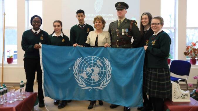 Schüler der Drogheda Grammar School und Personen in Militäruniform halten gemeinsam eine Flagge der Vereinten Nationen in einem Raum.