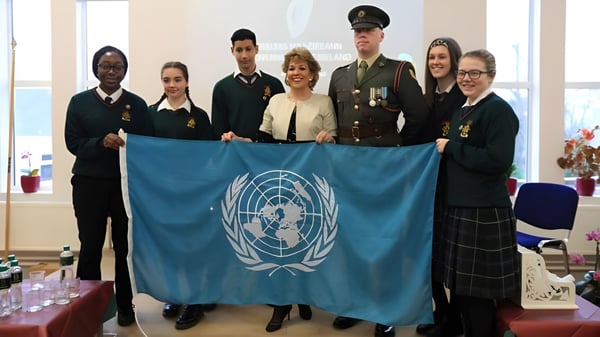 Schüler der Drogheda Grammar School und Personen in Militäruniformen halten gemeinsam eine Flagge der Vereinten Nationen in einem Innenraum.