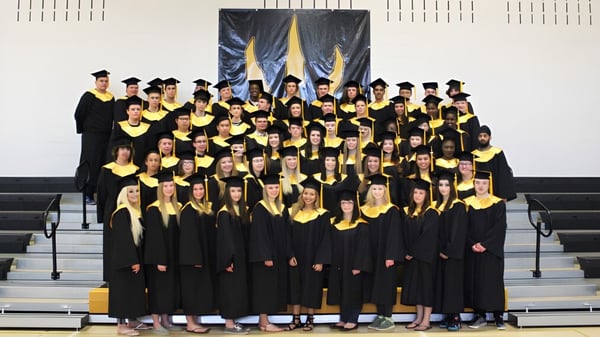 Eine große Gruppe von Schülerinnen und Schülern der Drumheller Valley School trägt schwarze Abschlusskleidung vor gelb-blauen Bannern.