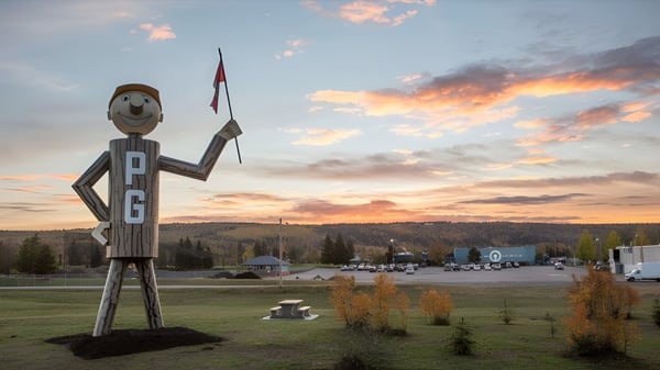 Eine große hölzerne Figur mit lächelndem Gesicht steht vor einer Landschaft mit Bergen und Bäumen auf dem Gelände der Duchess Park Secondary School.