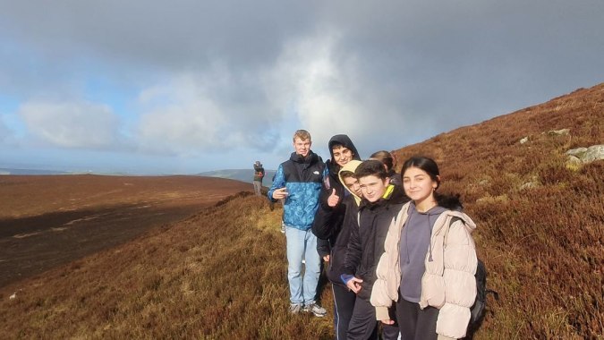 Eine Gruppe von Schülerinnen und Schülern steht auf einem grasbewachsenen Hügel mit bewölktem Himmel auf dem Gelände des Duiske College.