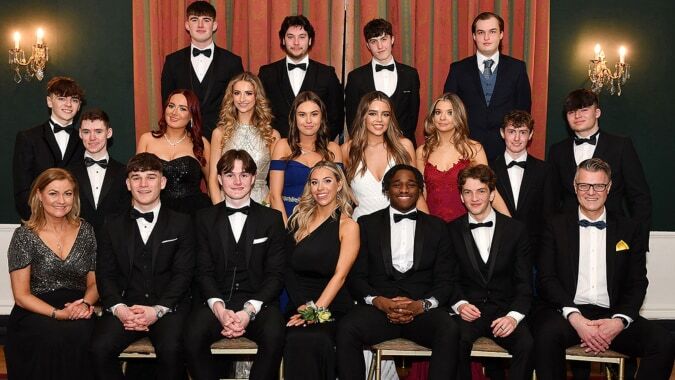 Eine Gruppe formell gekleideter Schülerinnen und Schüler der Dundalk Grammar School sitzt in einem festlichen Raum mit Kronleuchtern und Vorhängen.