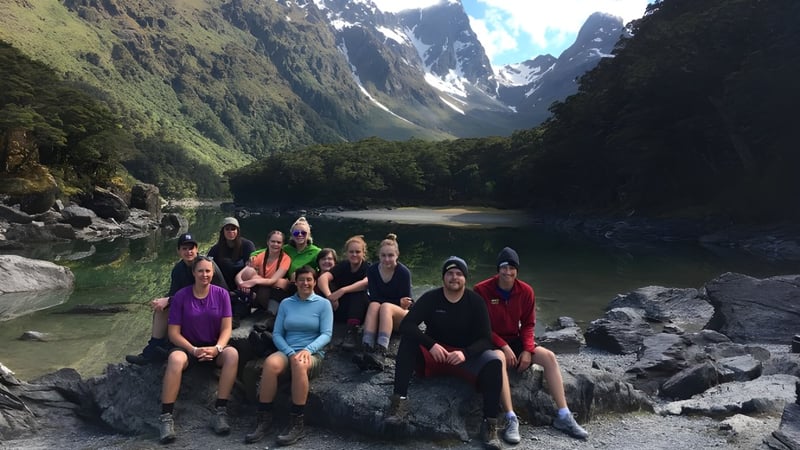 Eine Gruppe Schüler sitzt auf Felsen in einer Berglandschaft mit See und grüner Vegetation bei Dunstan High School.