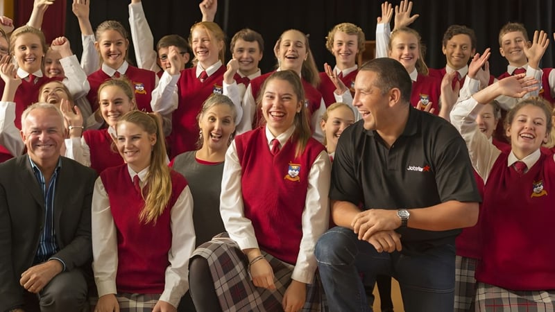 Eine Gruppe von Schülerinnen und Schülern in roten Uniformen sitzt und steht zusammen, daneben sitzt ein Mann im schwarzen Hemd, auf dem Campus der Dunstan High School.